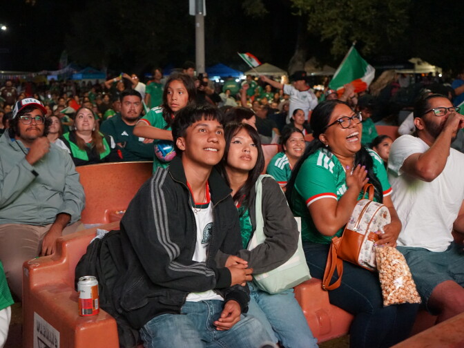 A crowd watches a World Cup match on a large outdoor screen. Many of them wear green jerseys. A young man and woman hold hands, sitting on a bench.