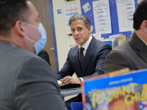 A man in a pinstripe suit sits at a table in a classroom, looking at other men in suits.