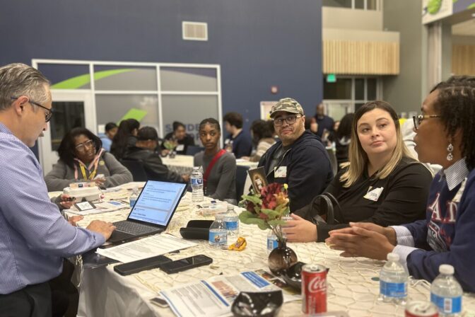 A group of men and women sit at a table, having a discussion. On the table are water bottles, papers, cellphones and a laptop