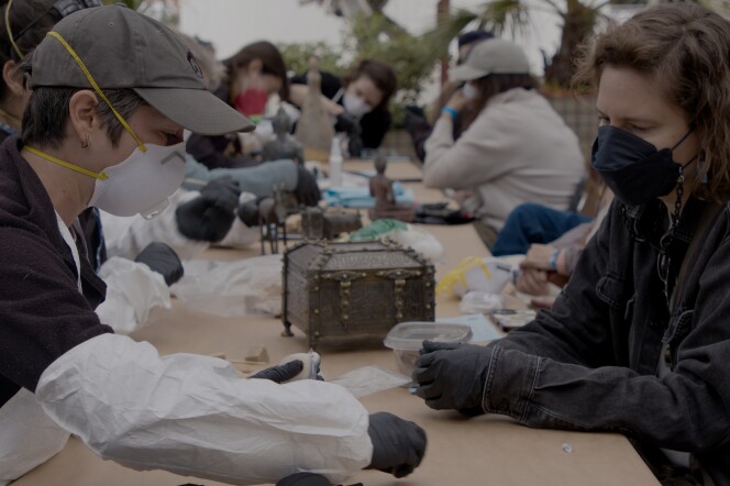 A conservator consults with a community member about their damaged object, assesses the condition, and proposes potential next steps. Both wear masks and sit across from each other at a table. 