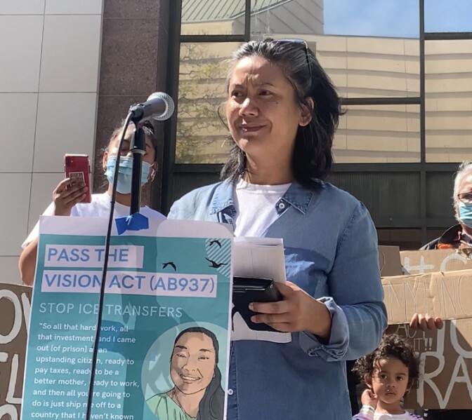 An Asian American woman in a denim shirt stands before a microphone with a sign taped to it that reads "Pass the Vision Act"