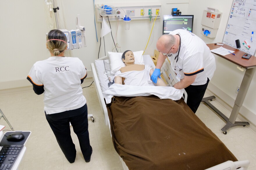 A man in a white uniform, bends over a hospital bed and examines a mannequin's arm. A woman stands next to the bed with her back to the camera. Her white uniform has the letters "RCC" on it.