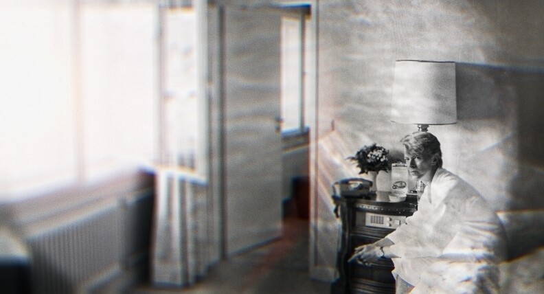 A black and white photo of David Bowie sitting on a bed in what looks like either an apartment or a hotel room, next to a stereo, background blurred and a wavy light filter over the entire image.