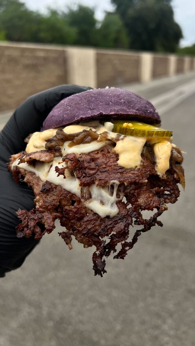 A hand holds a burger. The burger has a purple bun, crispy meat, melted cheese, pickles, and sauce.