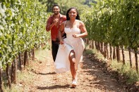 A female-presenting person in a white dress and a male-presenting person in an orange shirt running through a grove of young trees.