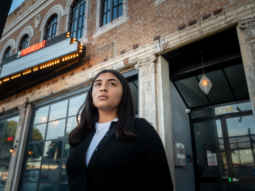 A young woman with dark hair and wearing a black sweater stands in front of a brick building with a sign that reads "The Paramount."