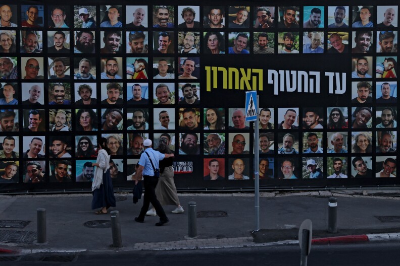 A large wall contains many photographs, along with Hebrew text. People walk past on a sidewalk.