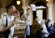 A man wearing a white hoodie, holds a brown poster that reads "KDL is Anti-Poor, KDL is Anti-Black, KDL is Anti-Native, VETO 41.18. He's holding his hand to his mouth as he shouts. Next two him a white woman holds up an out of focus white sign. On the right of the frame other diverse members of the public are standing, some holding signs up. 
