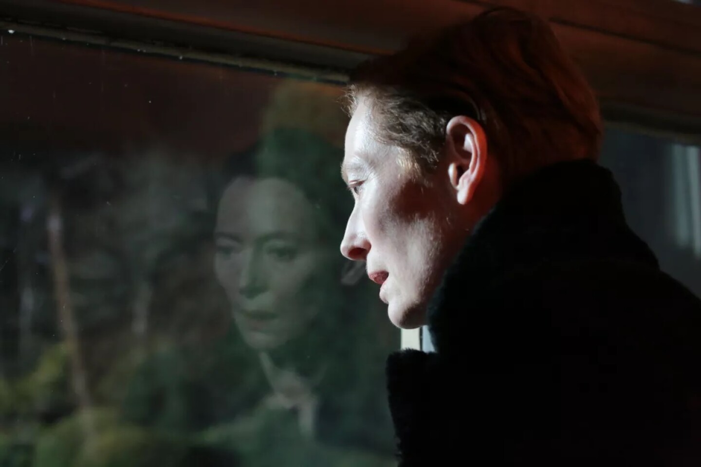 Tilda Swinton stands in front of a window right of frame. She can only be seen in profile as she stares out of the window. She wears a black coat with the collar pulled up to shield her neck. Her reflection can be seen in the window along with the green trees outside. The reflection shows her blank expression.