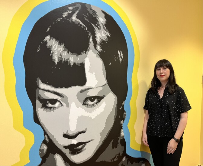 A woman of Chinese descent stands next to a large image of the 1920s silent screen star Anna May Wong, set against a bright yellow wall. 
