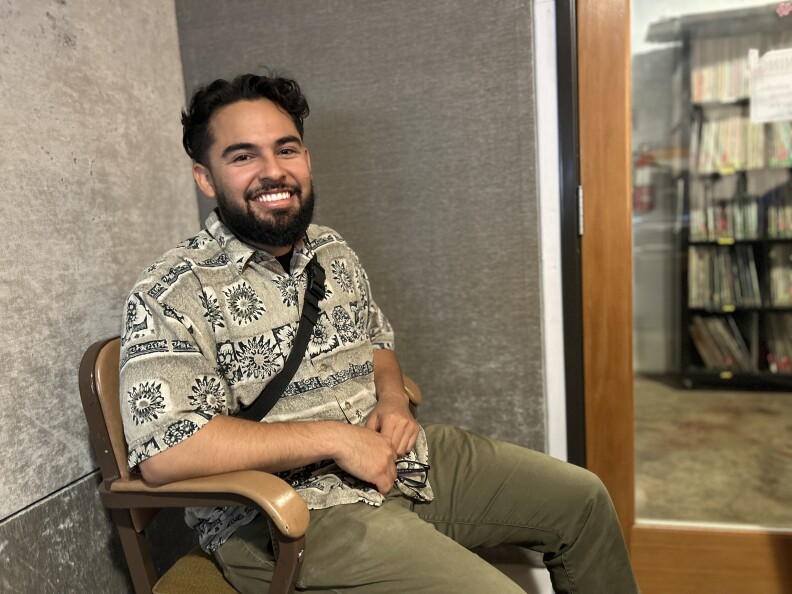 A man with light brown skin and a dark short hair and a beard, wearing a tan, patterned shirt and green pants sits in a a chair in silver velvet studio room, smiling. 