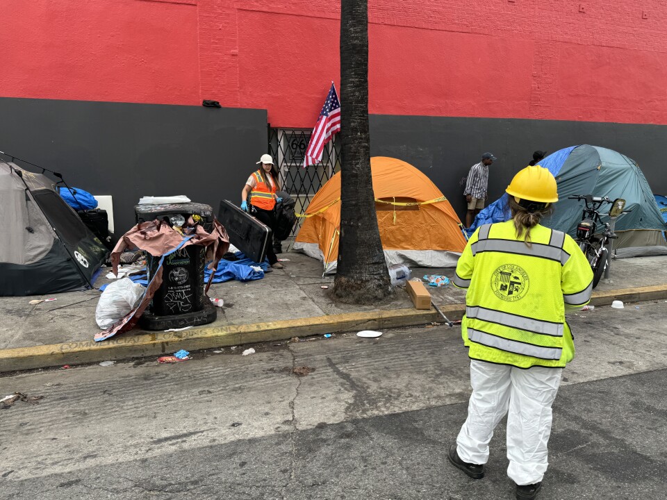A worker in a bright orange vest carries an object from between tents on a sidewalk, as another worker in a bright yellow vest looks on.
