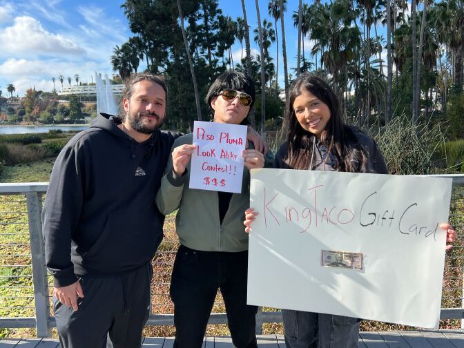 Three people at a city park. The one in the middle is holding up a paper reading "Peso Pluma Look Alike Contest!!! $$$"