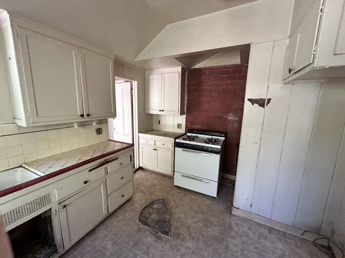 A kitchen inside a home, with an oven and sink. The vinyl flooring has a large damaged spot, and the white wall near the stove is chipped.