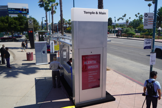 A person stands in the shade provided by a bus shelter as other people stand and walk around at a transit station next to a street.
