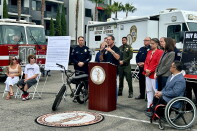 A man speaks at an Orange County District Attorney podium beside an e-bike, with officials and vehicles from fire, sheriff and DA departments behind him.