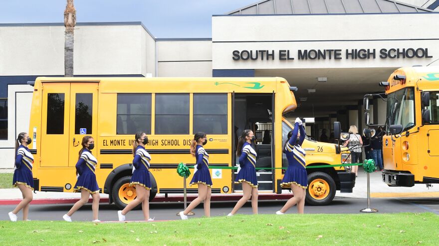 Cheerleaders in blue and gold uniforms walk in a single line past two bright yellow and black school buses. The buses are parked in front of a gray building that has the words "South El Monte High School" displayed in front.