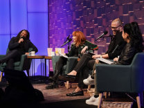 Four people are on a stage, sitting in green upholstered chairs with microphones in front of them. The person on the far left is a Black man wearing a black suit and shirt. To his left are 3 people: A red-headed white woman wearing a green and red track suit jacket and black pants; a biracial man with a shaved head and glasses; an Asian woman with long black hair in a ponytail. The last woman's face isn't visible. She is facing the other guests and host.