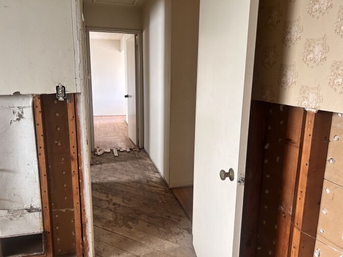 A hallway of a home, with the wooden flooring ripped and missing through most of it. Large portions of the wall on both the left and right hand side closest to the camera are also missing.
