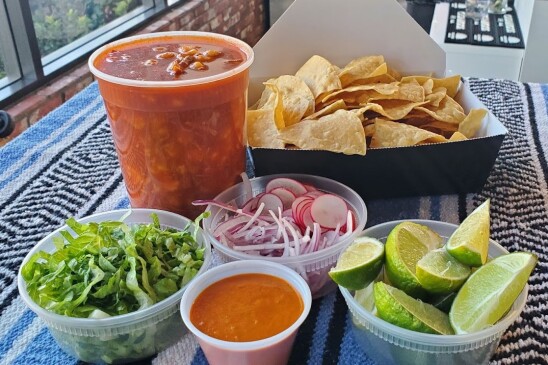 An image of pozole in a plastic container with tortilla chips, limes, salsa, radish, lettuce, on a table. 