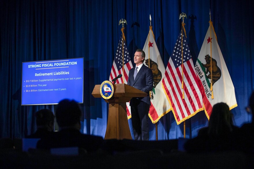 California Governor Gavin Newsom stands and speaks at a podium in front of two California flags and two American flags. He wears a navy suit and tie. A powerpoint presentation is shown on a large screen next to him.