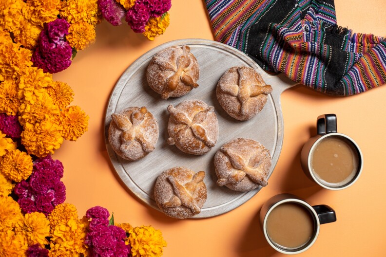 A tray of pan de muerto is accompanied by two cups of coffee and fresh marigold flowers.