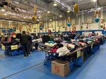 People peruse rows of tables laden with neatly-folded clothes. Mylar balloons are tied throughout the space. Some balloons are heart-shaped and some represent the letters "X," "M," "K," "S," and "L." They guide shoppers to tables with clothes that are their size. The tables are inside a basketball gym. 