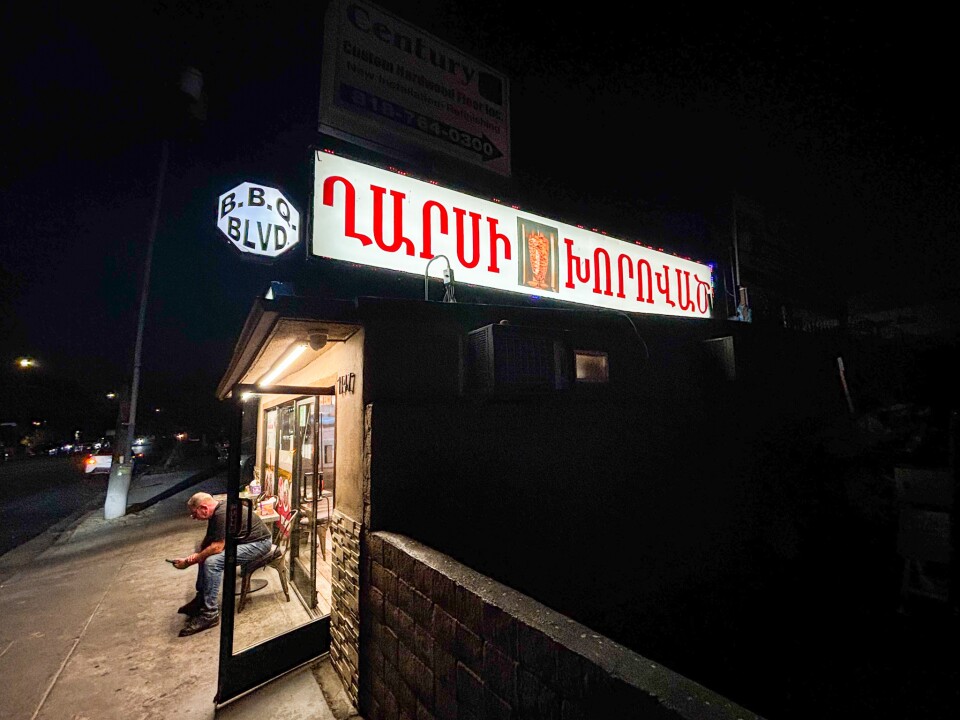 A small corner restaurant at night with a brightly lit sign in Armenian script and an image of shawarma meat. A man sits on a chair near the entrance under the glow of the lights, while a “B.B.Q BLVD” street sign hangs above.