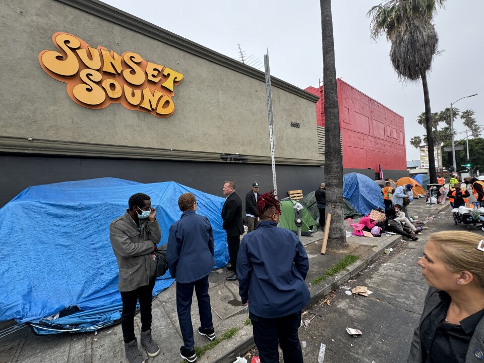 A group of people stand in front of tents on the sidewalk, in front of a building that says "Sunset Sound."