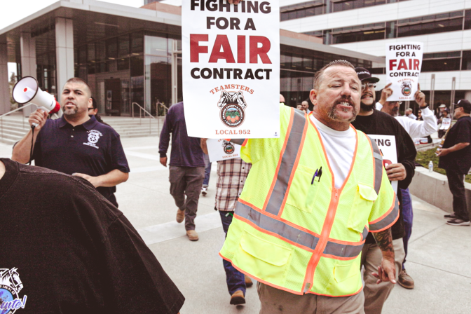 A man in a neon vest is walking and holding a sign that reads "For a Fair Contract Teamsters Local 952". Crowds of people behind him are also walking and holding signs.