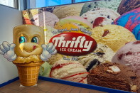 A life-sized cardboard cutout of Thrifty Ice Cream's smiling cone mascot giving two thumbs up, standing in front of a wall mural of colorful ice cream scoops and the Thrifty Ice Cream logo, "Since 1940."