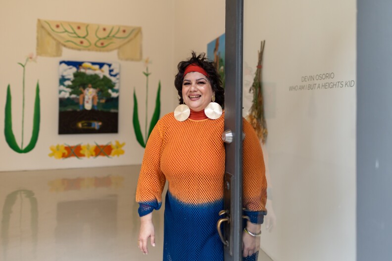 A woman, clad in a bright orange and blue dress with a red headband, poses for a photo. She smiles as she holds open the door. Artwork and drying flowers are in the background.