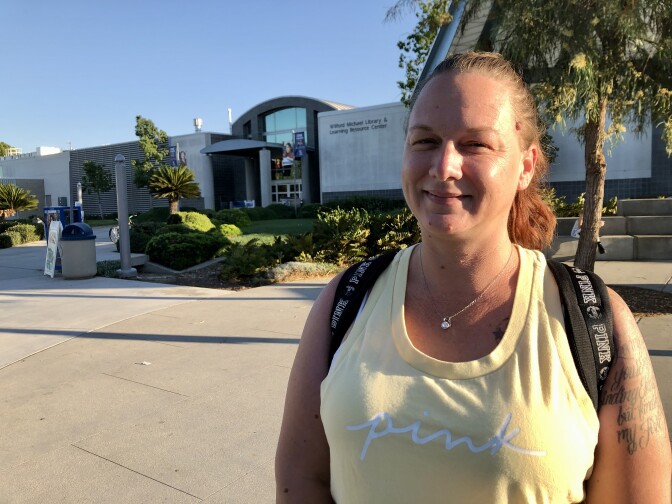 A light-skinned woman in a bright yellow tank top and a backpack smiles at the camera. In the background is a large college building.