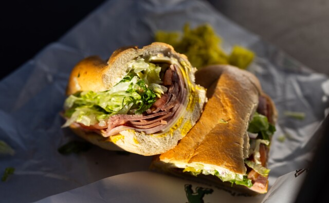 A close up, slightly overhead image of an Italian sandwich from Mickey's Deli in Hermosa Beach. The sandwich sits in two pieces and has been cut down the middle. The portion on the left sits on its side facing towards the camera, exposing the contents, which contains a pile of pink cold cuts, red tomatoes and green lettuce. The cut inside of the bread also shows a thin layer of yellow mustard on the bottom.