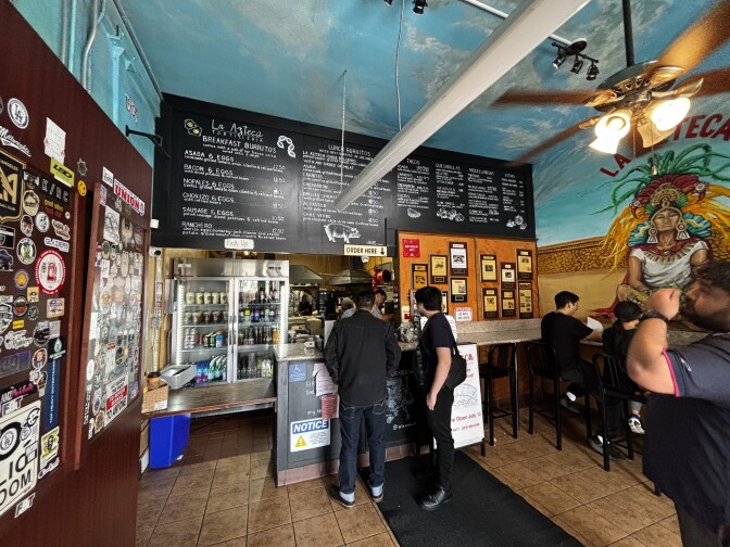 The interior of a restaurant with two people ordering at the counter. There's a large menu above, stickers over many of the walls, and murals showing Aztec culture.