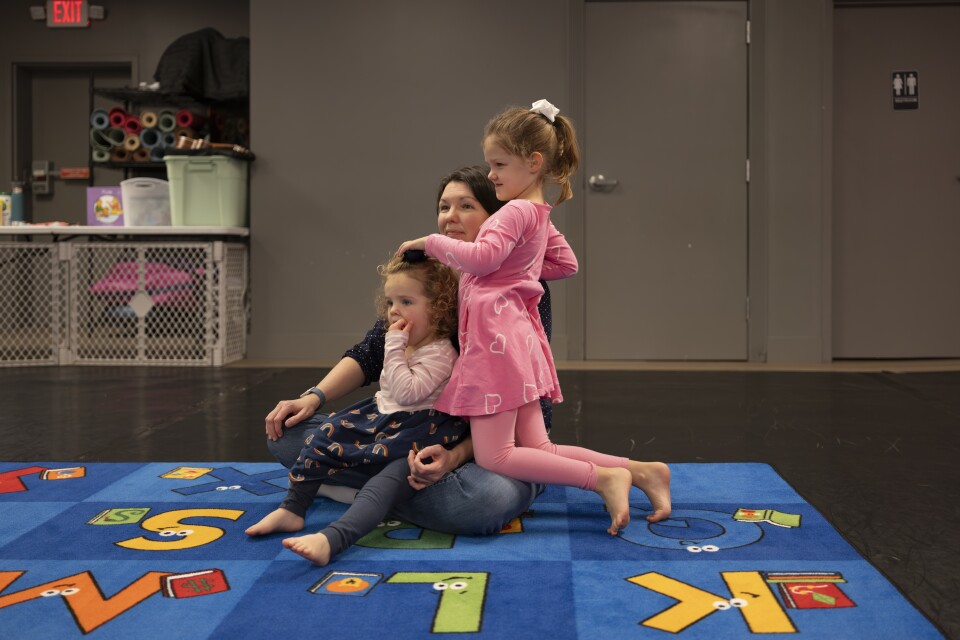 A woman and two young children with light skin tone sit on a carpet with an alphabet design.