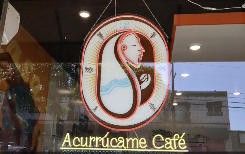 The front window of a store which says Acurrúcame Cafe in neon; above it is a side ways facing face surrounded by coffee symbols