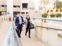 A young man in a suit and a middle-aged woman in a blazer walk up a ramp, both wearing expressions of tearful joy. 