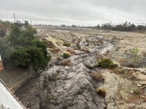 An overhead shot of an ashy muddy river rushing in a large dirt debris basin. The sky is overcast. 