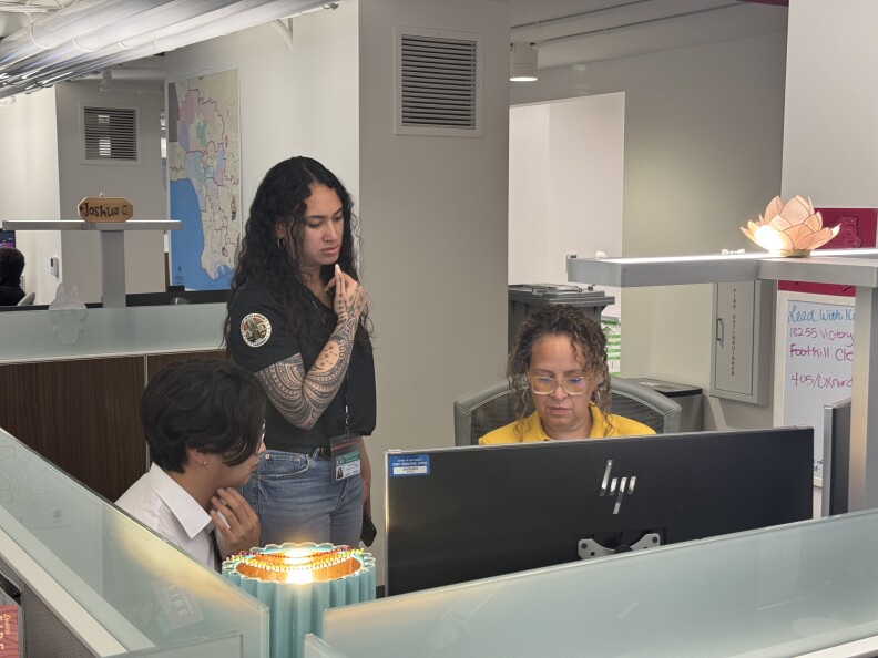 Three people in an office are facing a grey computer. One of the people is standing.