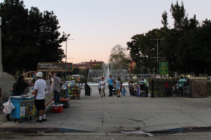 The Exposition Park Rose Garden was busy on a recent Thursday night. The fountain is spraying water. Kisd are walking towards the camera. Street vendors sell hot dogs, ice cream, and chips to the left. USC and the setting sun are in the background. 