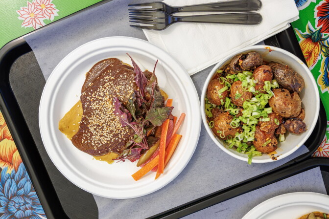 A banana-leaf-steamed tamal on a white plate, covered in dark brown mole and sprinkled with sesame seeds, with a small salad of red-purple amaranth leaves and sliced pickled carrots. A bowl of roasted potatoes topped with green onions sits beside it on a tray over a floral oilcloth.