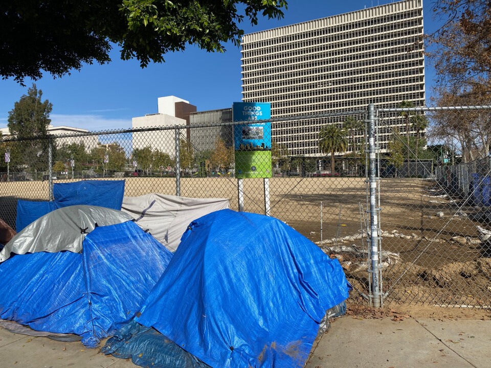 Two blue tents used by people experiencing homelessness are in front of a fence that is blocking off nearly 2 acres of dirt meant to be a park.