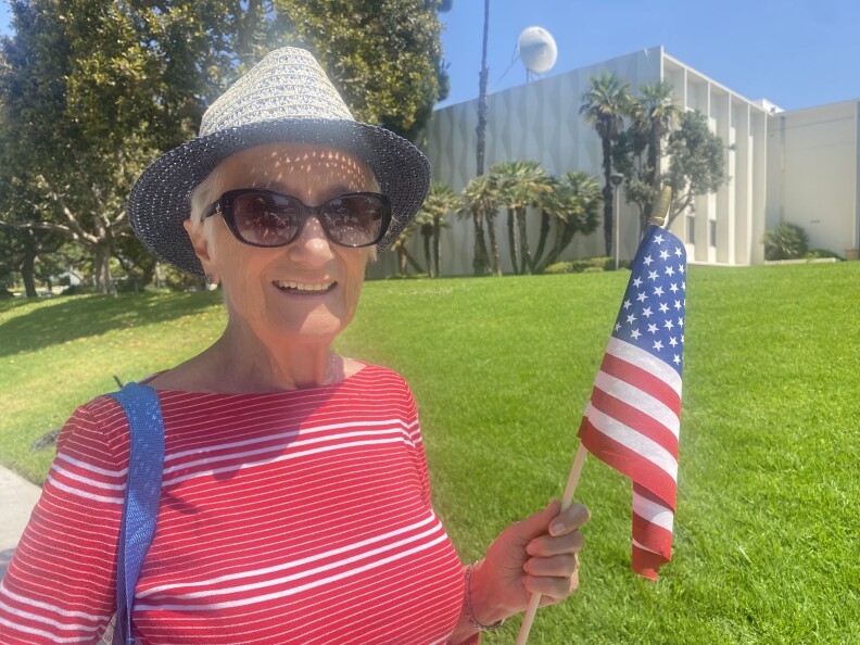 A woman, wearing a blue straw hat, a red and white striped top and sunglasses, is holding an American flag and smiling.