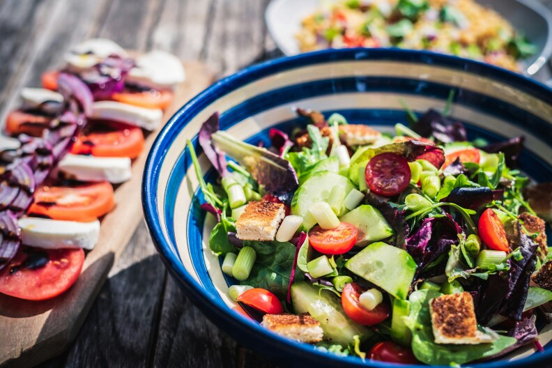 Fresh salad and caprese topped with red onions, displayed in the daytime on an outdoor wooden table.