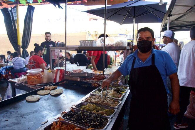 a man wearing a black apron and face mask stands next to a food cart where he is preparing tacos, taquitos and other items.