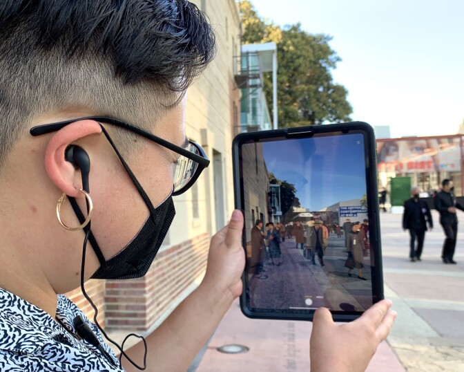 The side profile is visible of a person with short hair and hoop earrings, holding a digital tablet with an image of people in 1940s-era dress superimposed over the present-day scene of a courtyard.