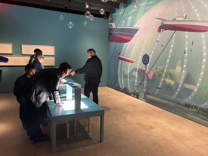 About four people are standing on the left side of two low desks, with one man standing on the other side on the right pointing towards one of the desks. A large mural of an underwater scene is painted on the back right wall.