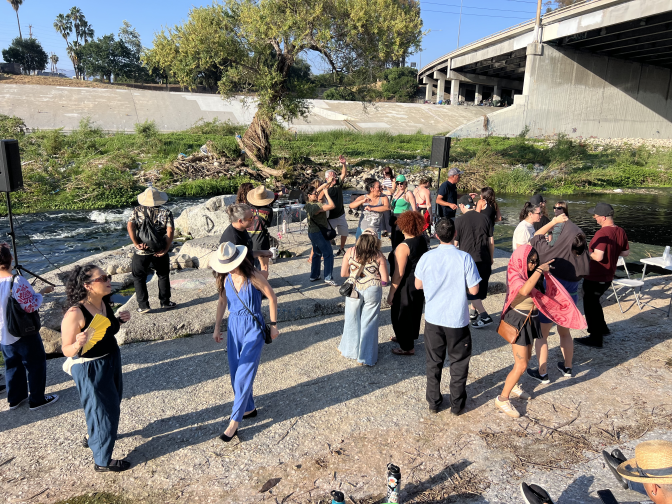 A group of people dancing in the concrete bed of the Los Angeles River is depicted. Speakers are seen on either side of the picture and a large tree is seen in the background.