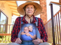 An older white woman holds an approximately 8 by 10 professional photo of herself and her husband smiling together at the camera. She sits outdoors on the steps of a porch and wears denim overalls, a red and blue checkered flannel shirt, and a straw sun hat with a black strap around her chin. 